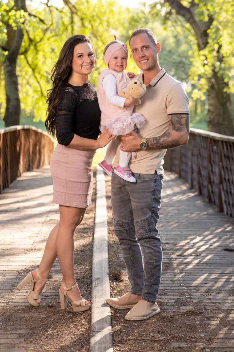 Családi fotózás Debrecen - Egy mosolygós család áll a híd közepén, a szülők büszkén tartják kislányukat, aki plüssállatot szorongat. A háttérben napfényben úszó, zöldellő fák emelik ki a pillanat harmóniáját.