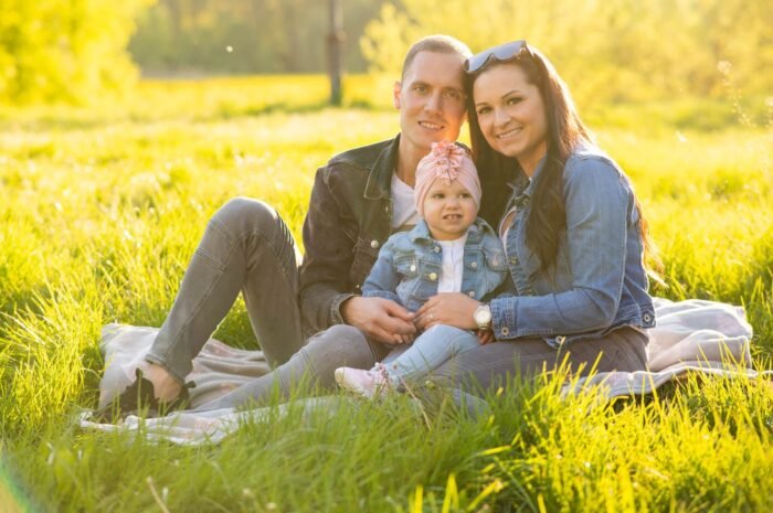 Szabadtéri családi fotózás. Egy mosolygós család ül egy pokrócon a zöldellő réten, a kislány középen helyezkedik el, szülei szeretettel ölelik. A napfény kellemes, aranyló ragyogással önti el a jelenetet, idilli hangulatot teremtve.
