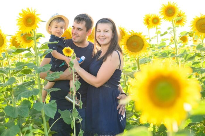 Egy fiatal család áll egy napraforgómezőben, mosolyogva néznek a kamerába. Az apa karjaiban tartja kisfiát, míg az anya mellettük áll, körülöttük aranyló napraforgók emelik ki a természet szépségét.