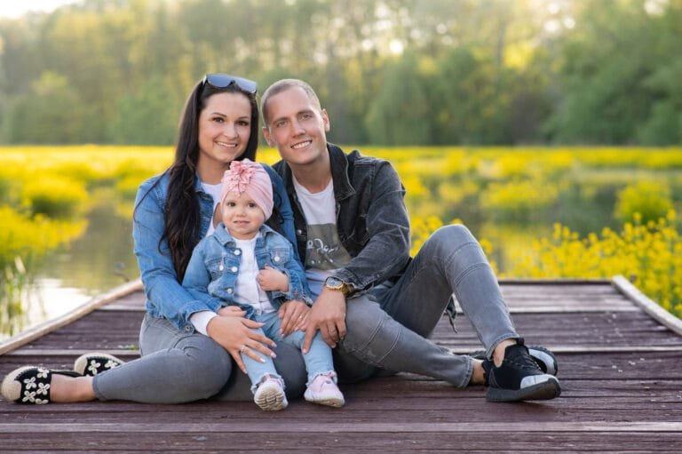 Családi fotózás Debrecen – egy család ül egy stégen a Vekeri-tónál, a szülők mosolyogva ölelik kislányukat. A háttérben sárgán virágzó növények és a tó tükröződése teremtenek békés, idilli hangulatot.