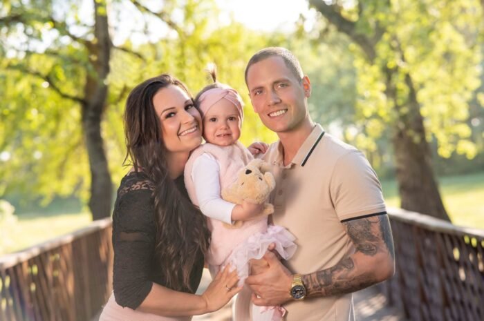 Szabadtéri családi fotózás Debrecenben. Egy boldog család mosolyog a kamerába, a kislányt középen tartják szeretettel. A háttérben napfényben fürdő zöld természet és egy híd fokozza a meghitt hangulatot.