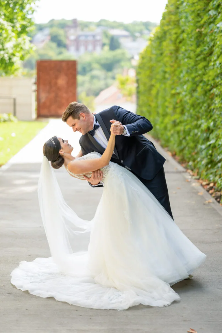 Esküvői fotózás Budapest szívében, a Várkert Bazár gyönyörű környezetében, ahol a pár egy romantikus tánccal ünnepli a szeretetüket. A menyasszony a vőlegény karjaiban, miközben a lenyűgöző zöld sétány és a távoli városképek mesés háttérként szolgálnak. A pillanat tökéletes megörökítése esküvői fotózáson.