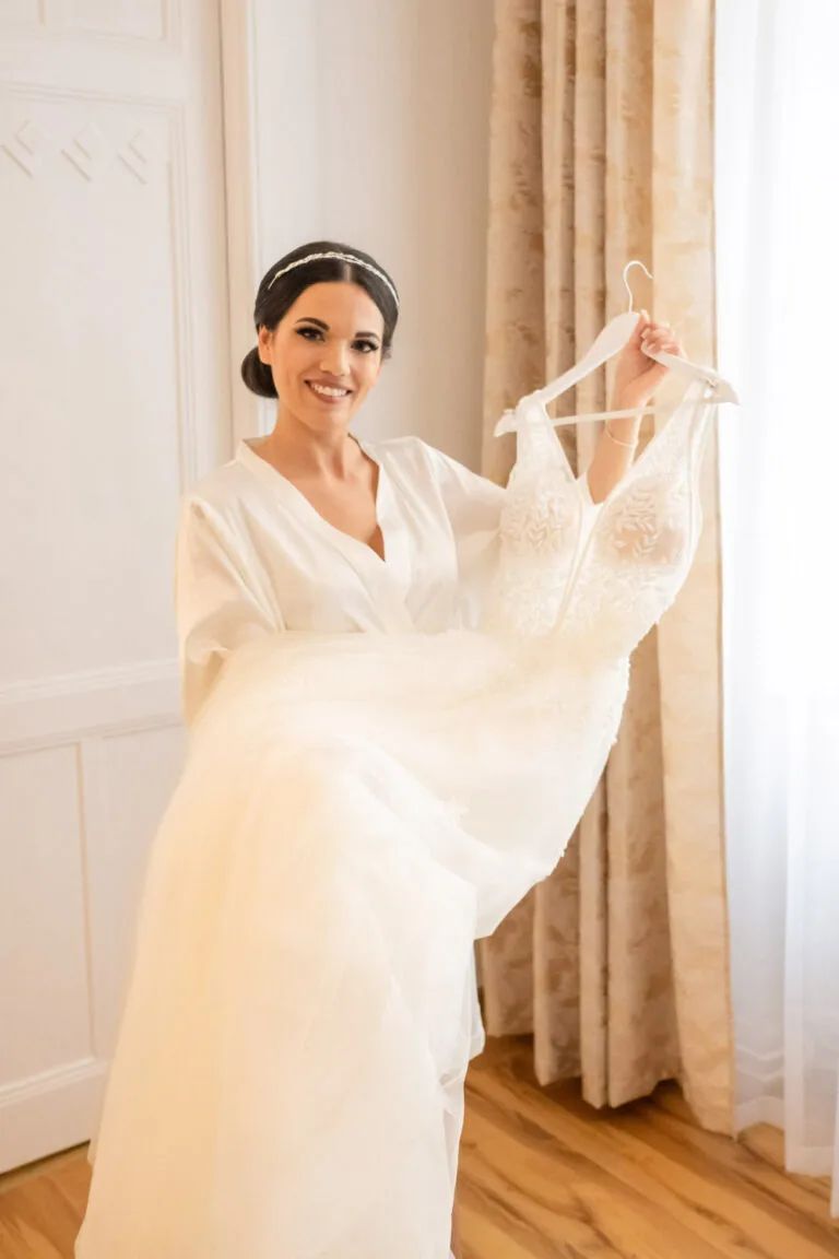 A menyasszony boldogan mutatja meg a gyönyörű esküvői ruháját, miközben készül a nagy napra. A természetes fényben, a háttérben lévő ablak által, a pillanat ragyogóan életre kel. Az esküvői fotós elkapja ezt a különleges pillanatot, amely örök emlék marad a menyasszony számára az esküvője előtti izgalmas készülődésből.