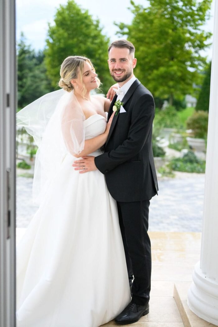 Az ifjú pár összbújva állnak, miközben a háttérben a természet romantikus színeivel díszíti a pillanatot.