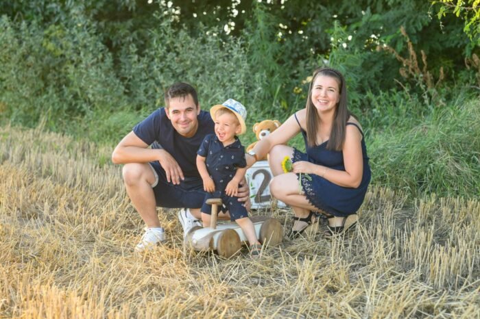 Egy mosolygós család ünnepli kisfiuk 2. születésnapját a szabadban. A kisfiú egy fa játékautón ül, mellette a szülők nevetve figyelik őt. A háttérben szalma és zöld növényzet teremtenek idilli hangulatot.