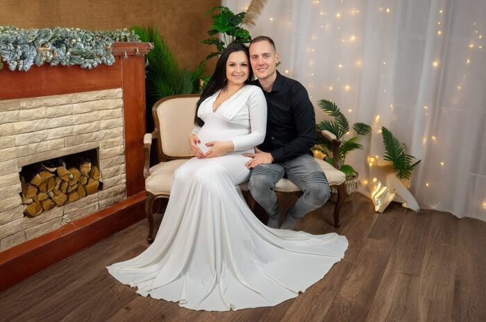 Egy mosolygós kismama és párja egy elegáns kanapén ül, ünnepi dekorációval körülvéve. A kismama fehér, földig érő ruhát visel, kiemelve a boldog várakozás pillanatait. A háttérben meghitt fények és zöld növények teszik különlegessé a fotózás hangulatát.