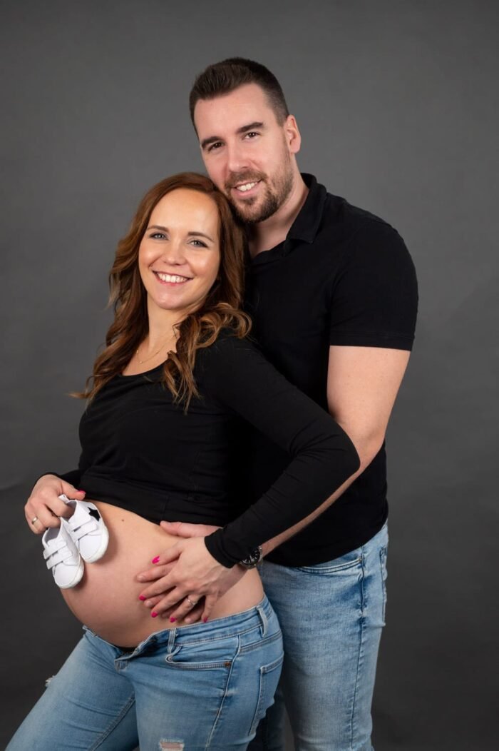 Kismama fotózás debreceni stúdióban, boldog pár mosolyog, a kismama kezében egy kis cipővel, fekete háttér előtt.