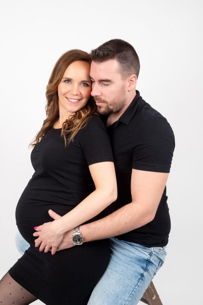Pocakos fotózás Debrecenben, boldog pár mosolyogva, a kismama fekete ruhában, párja szeretetteljesen öleli fehér háttér előtt.