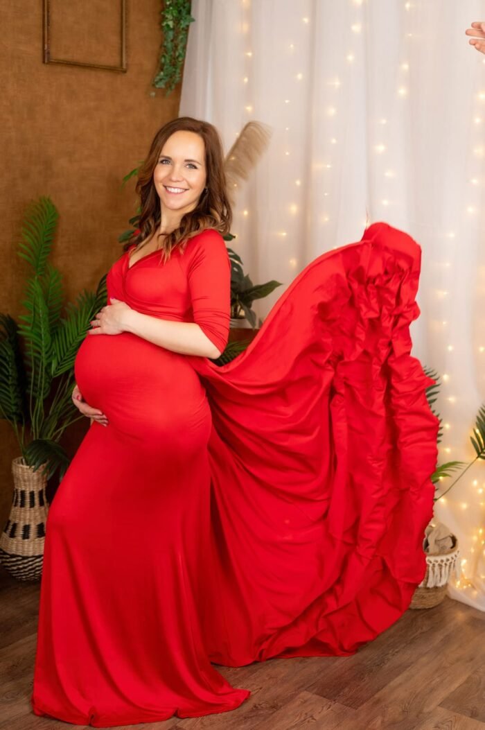 Kismama fotózás Debrecenben kismama ruhával- Kismama fotózás stúdióban, mosolygó kismama piros ruhában, romantikus hangulatú dekorációval és lebegő ruhaszoknyával.