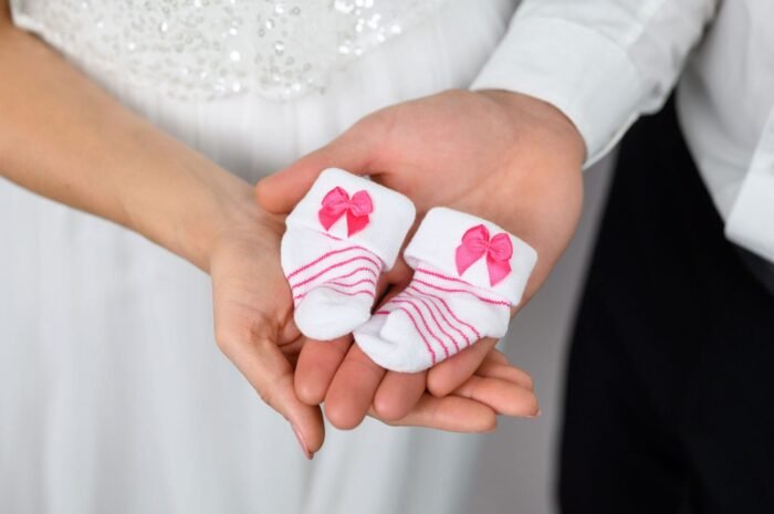 Közelkép két kézről, amelyek egy pár apró, rózsaszín masnival díszített babazoknit tartanak. A kép az új élet érkezésének meghitt és örömteli pillanatát szimbolizálja.