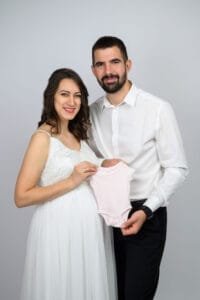 Kismama fotózás debrecen. Egy fiatal pár boldogan mosolyog a kamera felé. A kismama elegáns fehér ruhát visel, miközben egy kis babaruhát tartanak közösen, ünnepelve a családjuk gyarapodását.