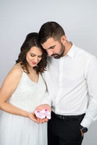 Egy fiatal pár szeretettel néz a kezükben tartott aprócska babazoknira. A kismama elegáns fehér ruhában van, a pillanat meghittsége pedig az új élet érkezésének örömét tükrözi.