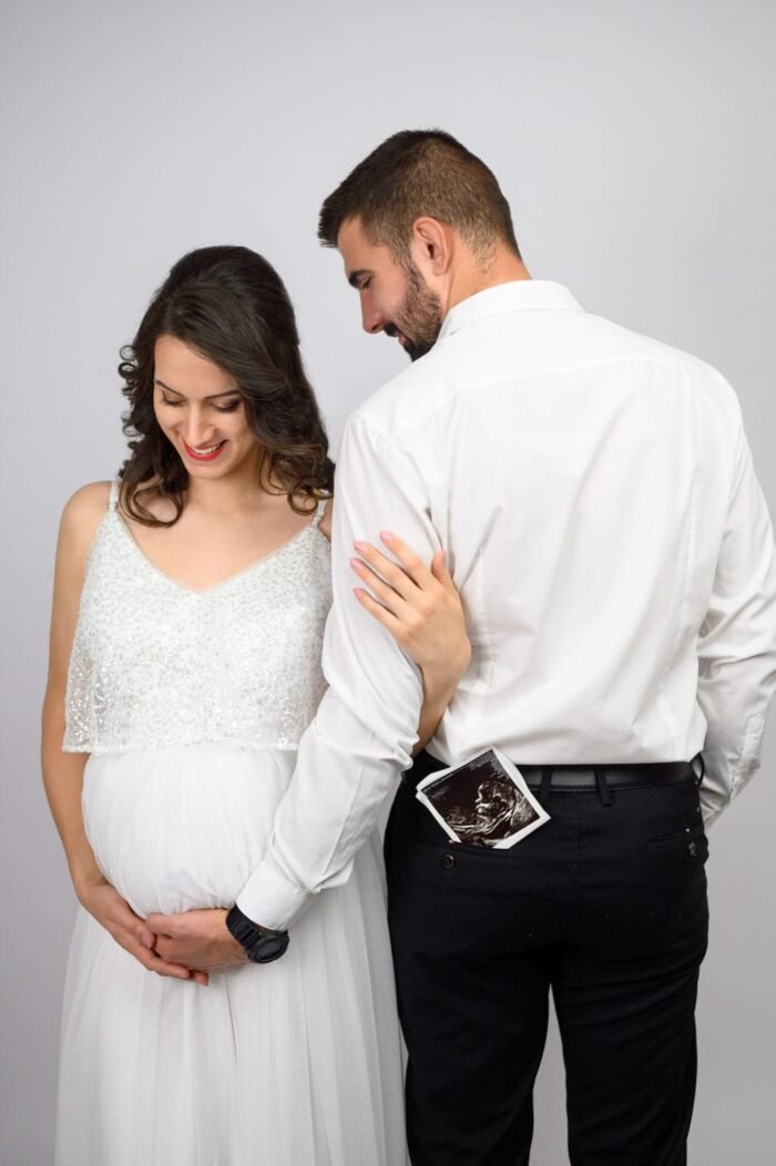 Kismama fotózás Debrecen - Várandós fotózás. A kismama gyengéden tartja a pocakját, miközben párja szeretettel átöleli őt. A férfi zsebéből egy ultrahang fotó látszik, amely a családalapítás boldogságát szimbolizálja.