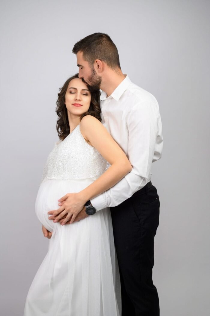 Egy boldog pár, ahol a kismama fehér, elegáns ruhában mosolyog, miközben párja szeretettel átöleli és homlokon csókolja. Meghitt pillanatot ábrázol a stúdió semleges háttere előtt.