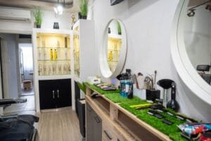 A barber shop belső terét bemutató kép, amely elegáns, modern tükröket, rendezett munkafelületet, hajápolási termékeket, valamint eszközöket és dekoratív növényeket tartalmaz. Az elegáns fekete székek és a tiszta, letisztult dizájn modern hangulatot áraszt.