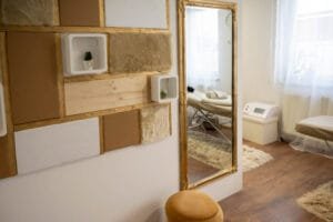 Kozmetikai szalon elegáns arany keretes tükörrel, modern dekorációs elemekkel és kényelmes masszázsággyal, amely ideális környezetet teremt a kikapcsolódáshoz és professzionális fotózáshoz.