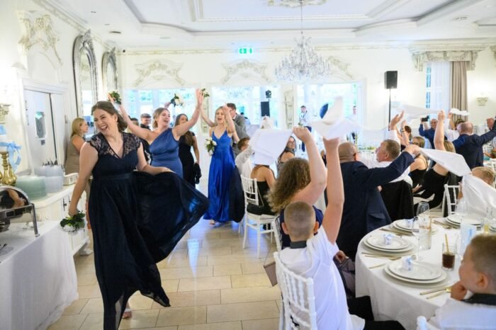 A koszorúslányok vidáman és elegánsan vonulnak be a díszterembe, miközben a vendégek fehér szalvétákat lengetve ünneplik az érkezésüket. A helyiség ragyogóan dekorált, a háttérben csillogó csillárok és elegáns tükrök láthatók.