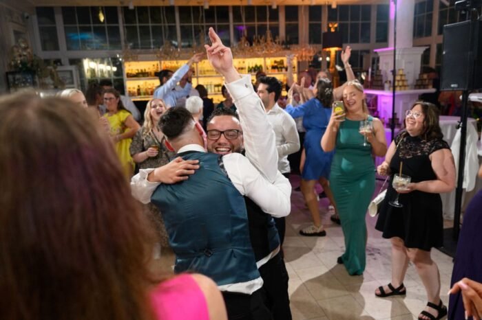 Az esküvői buli lendületes pillanata, ahol vőlegény barátai örömteli öleléssel ünnepel a táncparketten. A háttérben a vendégek táncolnak és nevetnek, miközben a vidám hangulatot színes fények és mosolygó arcok teszik felejthetetlenné.