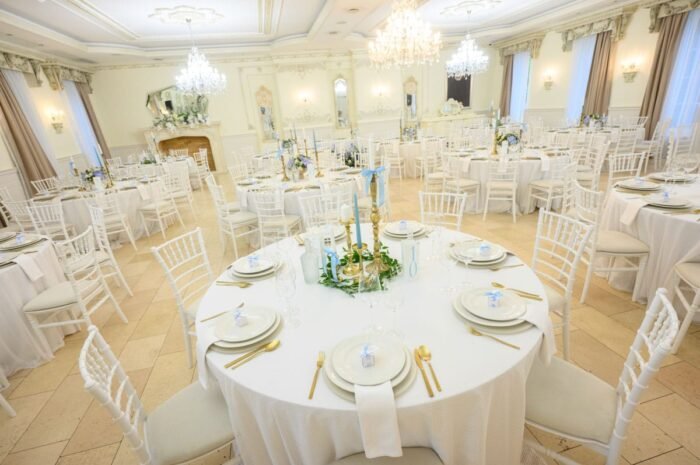 A Le Til Kúria elegánsan berendezett ünnepi díszterme, ahol a fehér terítők, arany evőeszközök és kék szalaggal díszített asztalok harmonikus hangulatot teremtenek. A csillárok ragyogása és a finom részletek emelik az esemény exkluzivitását.
