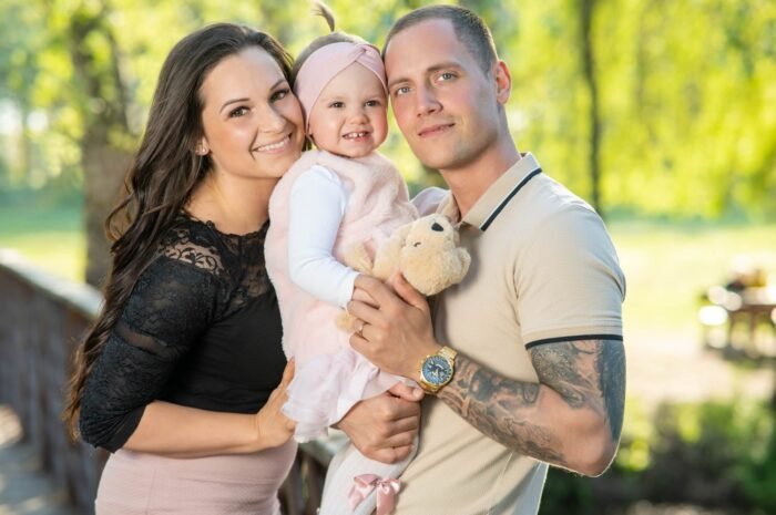 Egy boldog család mosolyog a kamerába, a kislányt középen tartják szorosan, miközben ő plüssállatát szorongatja. A háttérben természetes, zöld környezet teszi harmonikussá a pillanatot.