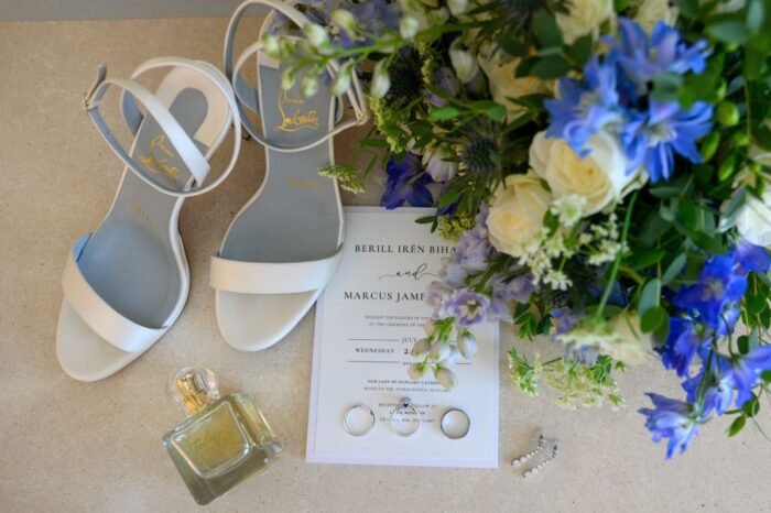 Esküvői részletek egy asztalon elrendezve: elegáns fehér szandálok, illatos parfüm, gyönyörű fehér és kék virágcsokor, esküvői meghívó, gyűrűk és csillogó fülbevalók. A kompozíció az esemény kifinomultságát és stílusát tükrözi.