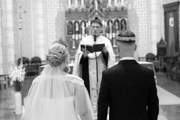 A menyasszony és a vőlegény a templomban állnak, koronával a fejükön, miközben a pap szertartást vezet. A fekete-fehér fotó kiemeli a pillanat méltóságát és spirituális jelentőségét.
