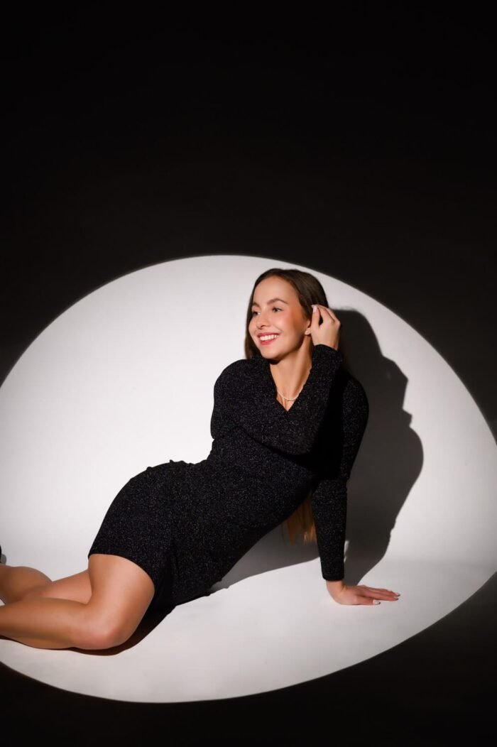 Egy fiatal nő fekete csillogó ruhában mosolyogva pózol stúdióban, fókuszált körvilágításban. A sötét háttér és a világos kör alakú fény kiemeli az elegáns megjelenését és a vidám hangulatot.