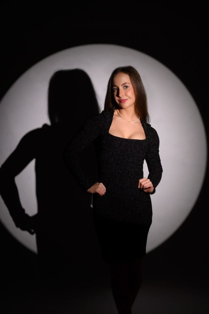 Egy fiatal nő fekete csillogó ruhában áll, miközben a kör alakú stúdióvilágítás kiemeli alakját a sötét háttérből. A nő kecses testtartása és mosolya elegáns megjelenést kölcsönöz a portrénak, miközben árnyéka érdekes vizuális elemet hoz létre.