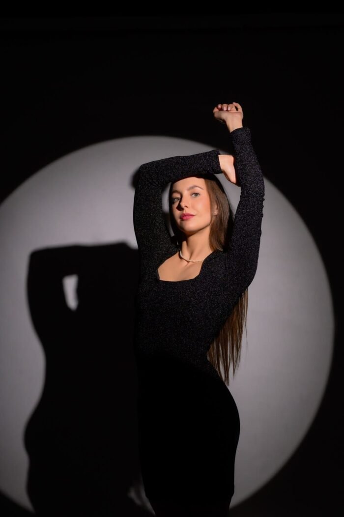 Portré fotózás Debrecenben - Egy fiatal nő fekete ruhában, kreatív pózban áll a kör alakú stúdióvilágítás középpontjában. Az árnyékok és fények játéka fokozza a portré dinamikáját, miközben a nő magabiztos arckifejezése és testtartása eleganciát sugároz.