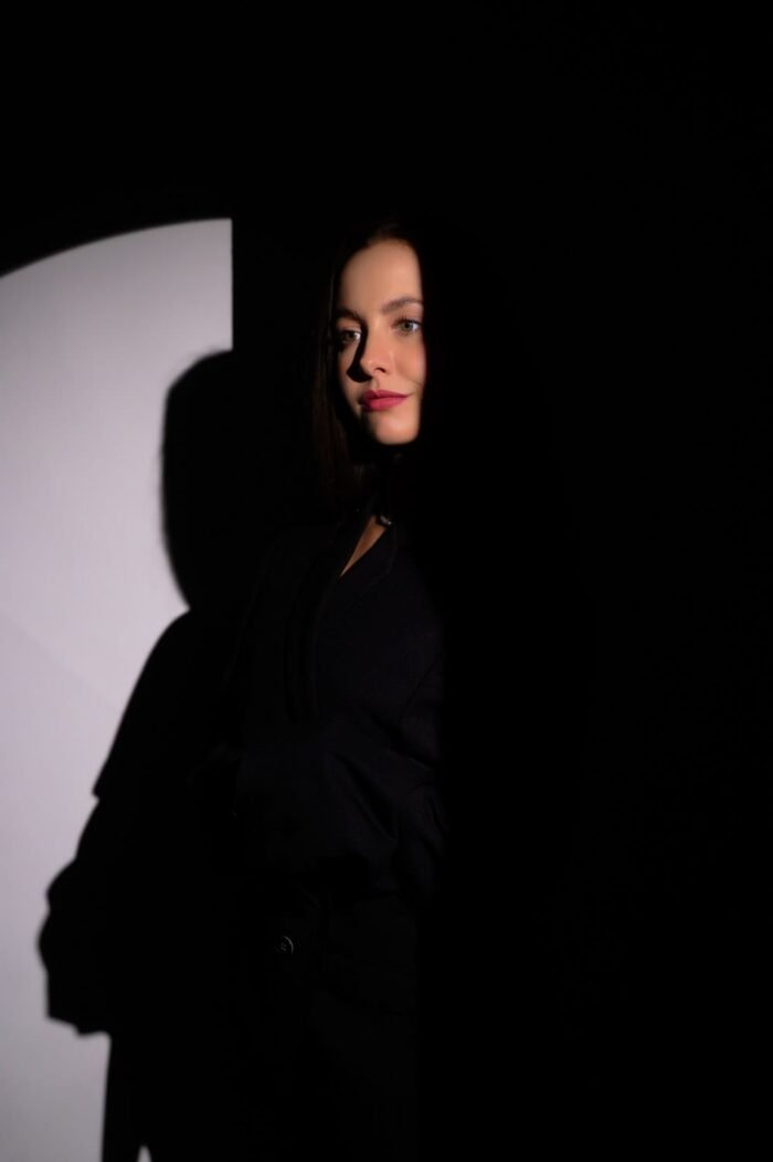 Egy fiatal nő arca részben megvilágítva jelenik meg a sötét háttér előtt, kiemelve a portré drámai és rejtélyes hangulatát. Az árnyék és a fény kontrasztja különleges atmoszférát teremt a képen.