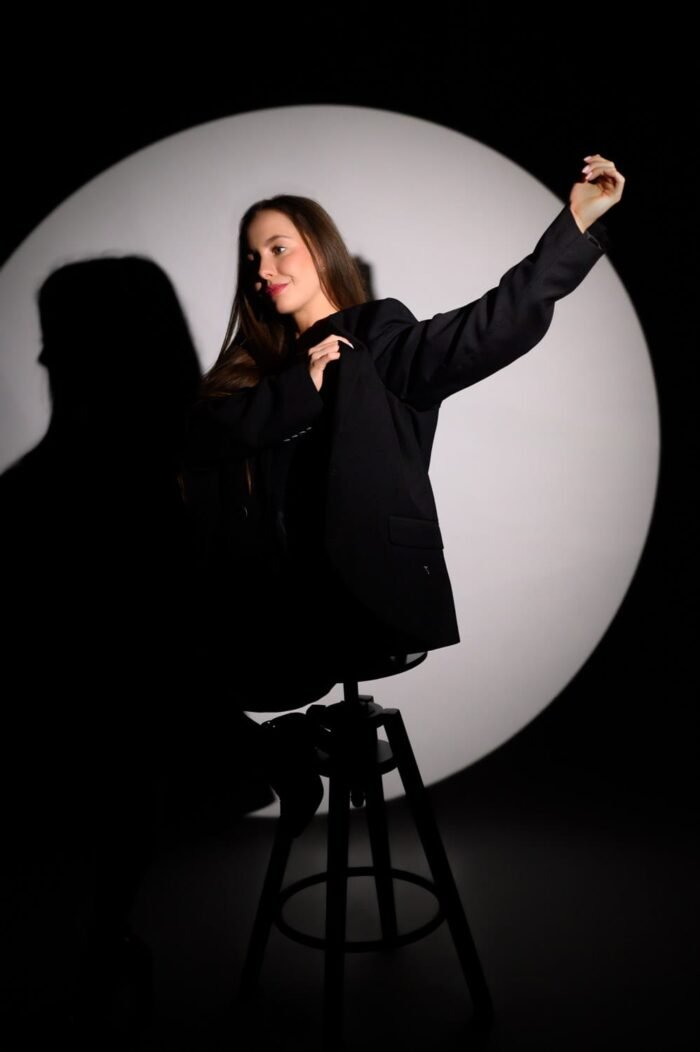Debreceni portré fotós - Egy fiatal nő fekete oversized zakóban ül egy stílusos széken, miközben egy játékos mozdulatot tesz a karjával. A háttérben kör alakú stúdióvilágítás emeli ki a sziluettjét, a sötét háttér pedig hangsúlyozza a kompozíció kreatív hatását.