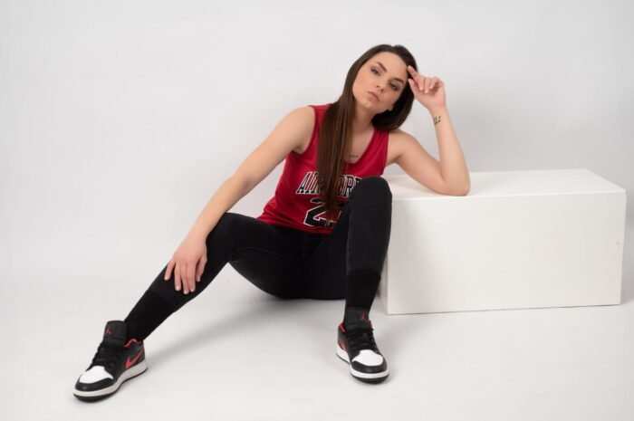 Egy fiatal nő piros Air Jordan trikóban, fekete nadrágban és fekete-fehér sportcipőben ül, miközben lazán tartja a kezét a fején. A kép letisztult háttérrel és stúdióvilágítással készült, mely kiemeli a sportos, mégis elegáns megjelenést.