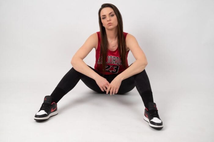 Egy fiatal nő sportos, kosaras ruhában, fekete nadrágban és Nike Jordan cipőben ül, a háttérben egy semleges fehér fal.