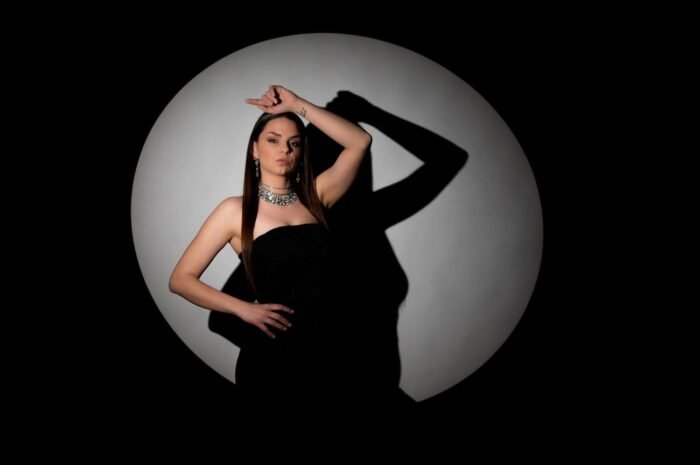 Egy nő elegáns fekete estélyiben, fényes nyaklánccal, háttérben nagy fénykörrel, amelynek árnyéka kiemeli a modell mozdulatait és kontúrját.