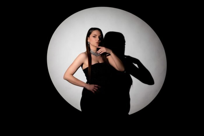 Elegáns fekete ruhás nő, csillogó nyaklánccal és fülbevalóval, pózol a reflektorfényben, amely kerek árnyékot vet a háttérre, kiemelve a fotó drámai hangulatát.