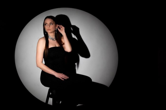 Egy elegáns nő fekete estélyiben, csillogó ékszerekkel pózol egy széken, háttérben kerek fény-árnyék kontraszttal, amely kiemeli az időtlen stílust és a finom részleteket.