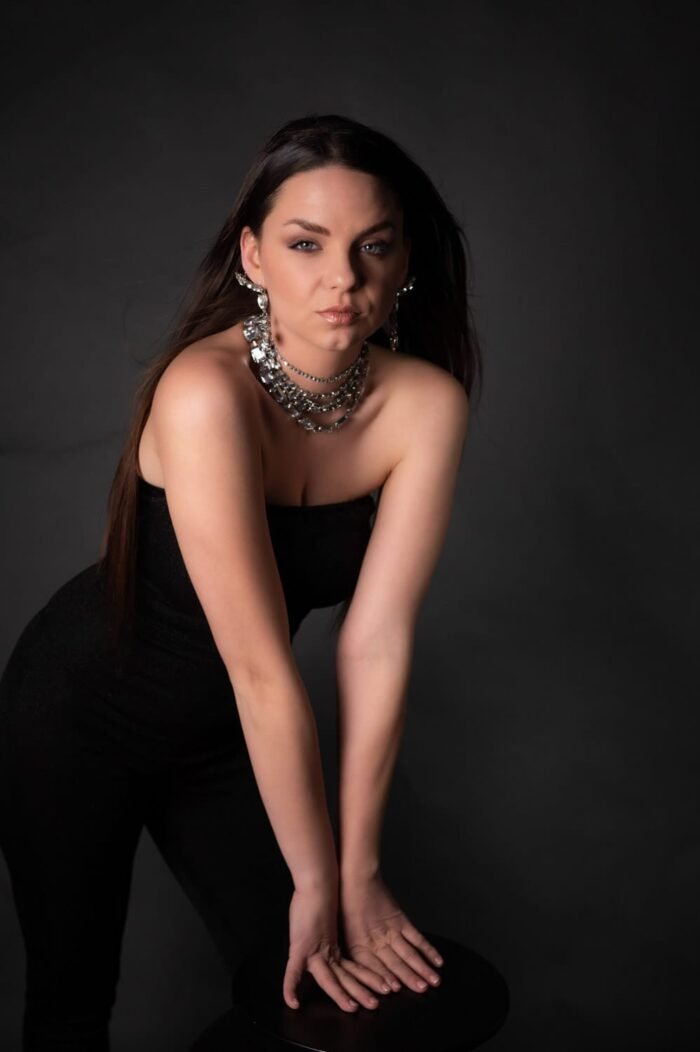Porté fotózás Debrecenben. Fiatal nő fekete ruhában, csillogó nyaklánccal és fülbevalókkal áll, kezeivel egy székre támaszkodva, klasszikus fekete háttér előtt, mely kiemeli az időtlen eleganciát és a tekintet magabiztosságát.