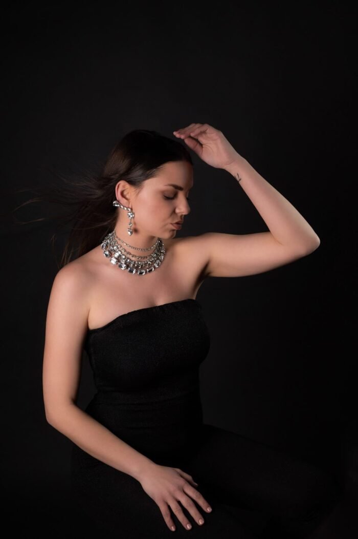 Modell elegáns fekete ruhában, csillogó ékszerekkel ül egy fekete háttér előtt. Szélfútta haja és finom, fejét érintő mozdulata időtlen eleganciát sugall.
