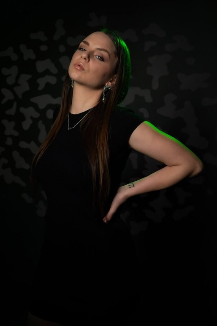 Modell fotózás Debrecenben. Egy fiatal nő fekete ruhában, elegáns ékszerekkel pózol, zöld fényben megvilágítva. A háttér geometrikus mintáival modern és különleges hangulatot teremt.