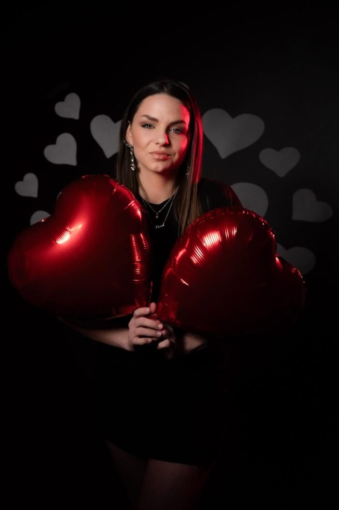 Fiatal nő fekete ruhában, elegáns ékszerekkel és piros szív alakú lufikkal pózol romantikus szívmintás háttér előtt, Debrecenben készült fotón.
