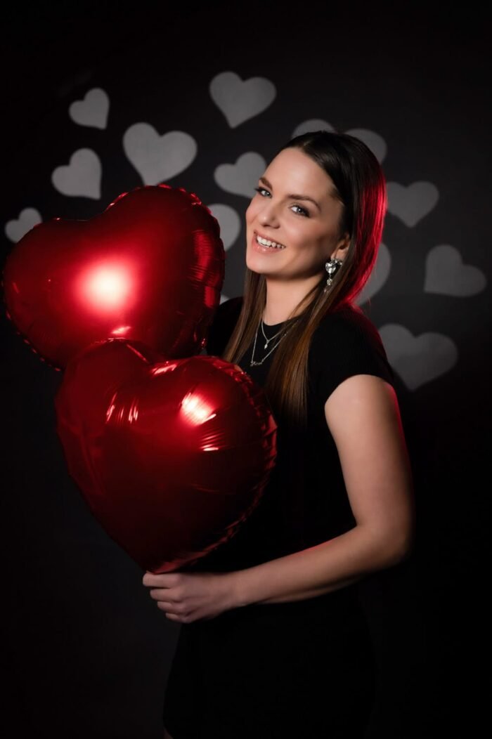 Ramóna fekete ruhában, elegáns ékszerekkel és piros szív alakú lufikkal pózol romantikus szívmintás háttér előtt, Debreceni stúdióban készült fotón.