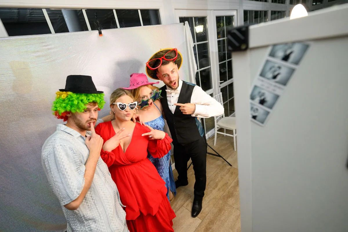 Vidám vendégek színes jelmezekben és vicces kiegészítőkkel pózolnak a selfiebox előtt egy esküvői bulin. Szelfigép bérlés Debrecenben