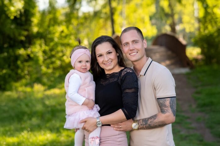 Családi fotózás Debrecenben - Egy mosolygós család áll a természetben, a szülők szeretettel ölelik kislányukat, aki rózsaszín ruhában és hajpántban tündököl. A háttérben zöld fák és egy híd fokozzák a meghitt hangulatot.