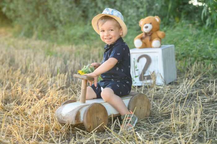 Gyermek szülinapi fotózás Debrecenben. Egy kisfiú ül egy fa játékautón a szabadban, kezében egy napraforgóval. A háttérben egy plüssmackó és egy "2" számot ábrázoló dekoráció jelzi a születésnapot, miközben a természet nyugodt szépsége keretezi a jelenetet.