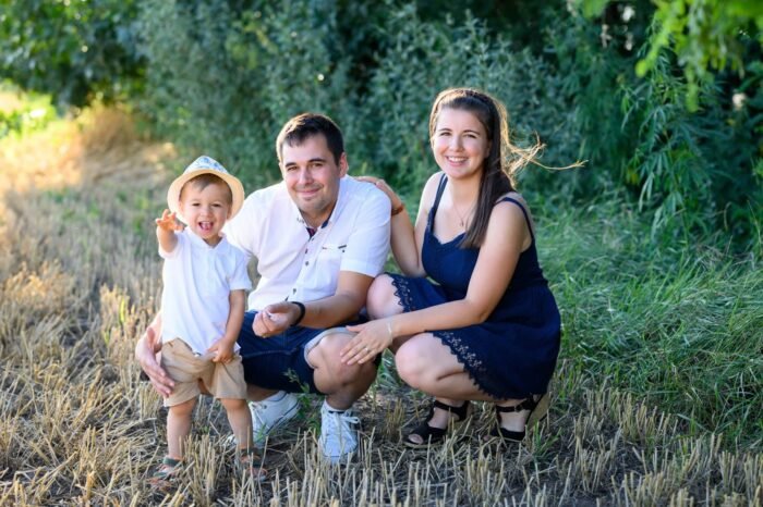 Szabadtéri szülinapi fotózás Debrecenben. Egy boldog család együtt ünnepli kisfiuk születésnapját a szabadban. A kisfiú mosolyogva integet, míg szülei szeretettel figyelik őt a háttérben zöld növényzettel és aranyló szalmával keretezve a jelenetet.