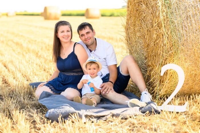 Szabadtéri családi fotózás Debrecenben. Egy mosolygós család ül egy szalmabála mellett a szántóföldön, miközben kisfiuk a 2. születésnapját ünnepli. A dekorációban szerepel egy "2" szám, és a háttérben aranyló szalmabálák láthatók, amelyek harmonikus vidéki hangulatot teremtenek.