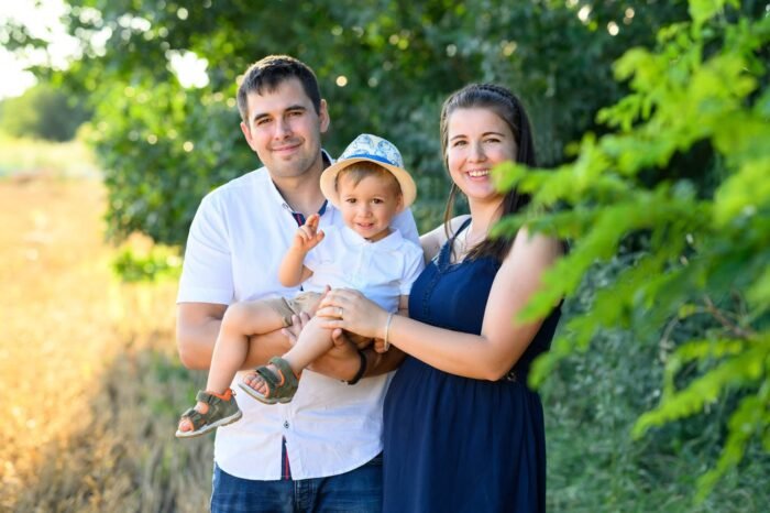 Egy mosolygós család ünnepli kisfiuk születésnapját a természetben. Az apa karjában tartja a kisfiút, aki kalapot visel és vidáman integet, miközben az anya szeretettel áll mellettük. A háttérben zöld növényzet és aranyló szántóföld teremt harmóniát.