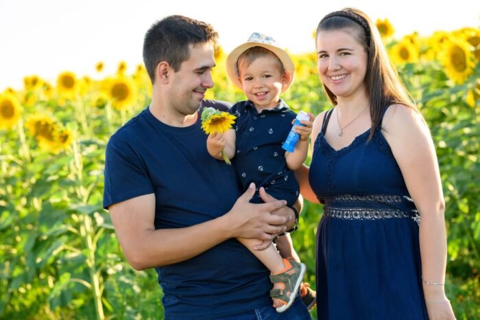 Egy mosolygós család ünnepli kisfiuk születésnapját egy napraforgómezőben. Az apa karjában tartja a kisfiút, aki egy napraforgót és egy buborékfújót tart a kezében, miközben az anya boldogan áll mellettük. A háttérben sárga napraforgók teszik színessé és vidámmá a jelenetet.