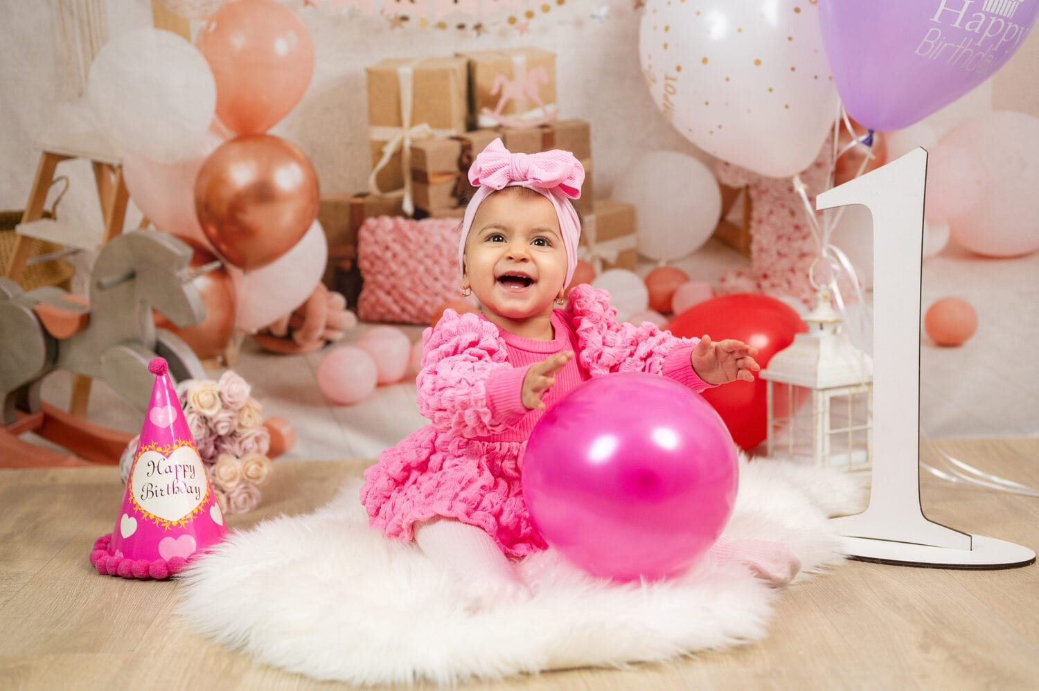 Egy éves szülinapi fotózás Debrecenben – Mosolygós kisbaba rózsaszín ruhában és masnival a fején ül egy fehér szőnyegen, miközben egy pink lufival játszik. A háttérben ajándékdobozok, lufik és egy fa hintaló teremtenek ünnepi hangulatot. Az első születésnap megörökítése profi stúdióban tökéletes emlék a család számára.