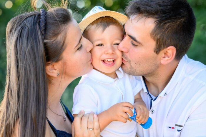 Debreceni fotós képe. Egy mosolygós kisfiút ölelnek szülei, miközben mindketten egy-egy puszit adnak az arcára. A kisfiú boldogan nevet, kezében egy buborékfújóval, a háttérben természetes, zöld környezet látható.